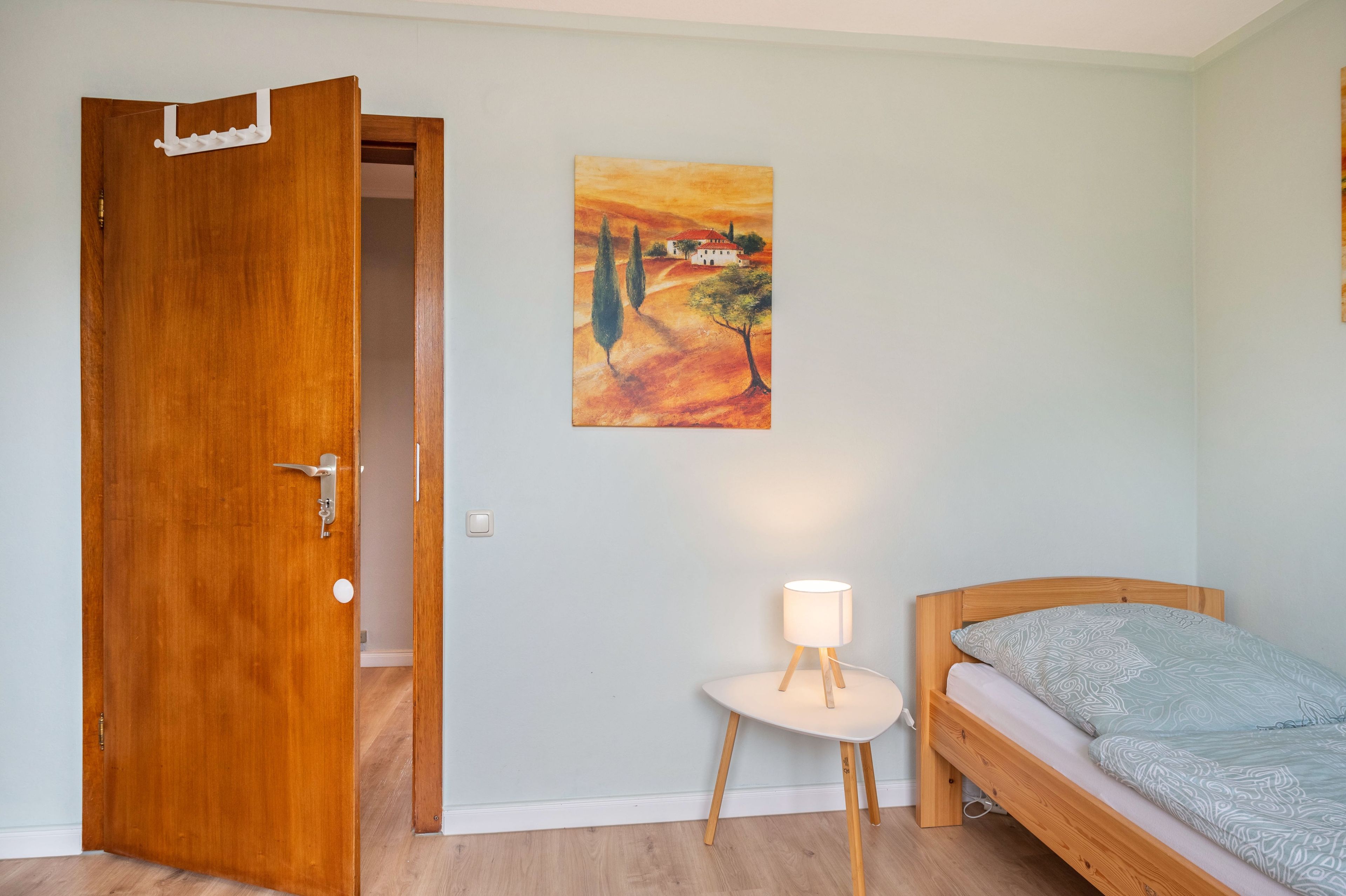 A room with a wooden door, a small bedside table with a lamp, a bed with patterned bedding, and a painting on the wall.