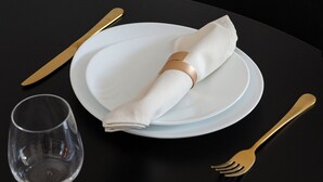 Ein Tisch ist gedeckt mit weißen Tellern, einem gefalteten Servietten, einem klaren Weinglas und goldfarbenem Besteck.