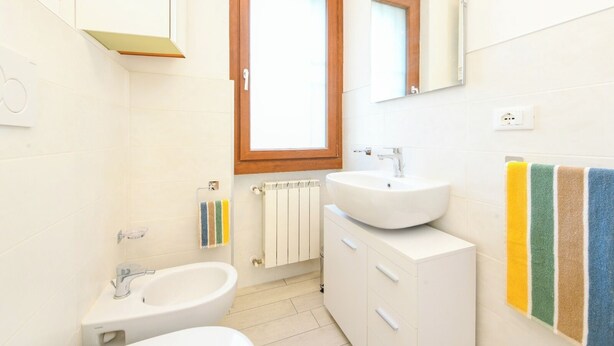 A modern bathroom with a bidet, a sink, and a mirror. There is a window above the sink and a radiator below it. - Sporting A12 by Wonderful Italy (Sirmione)