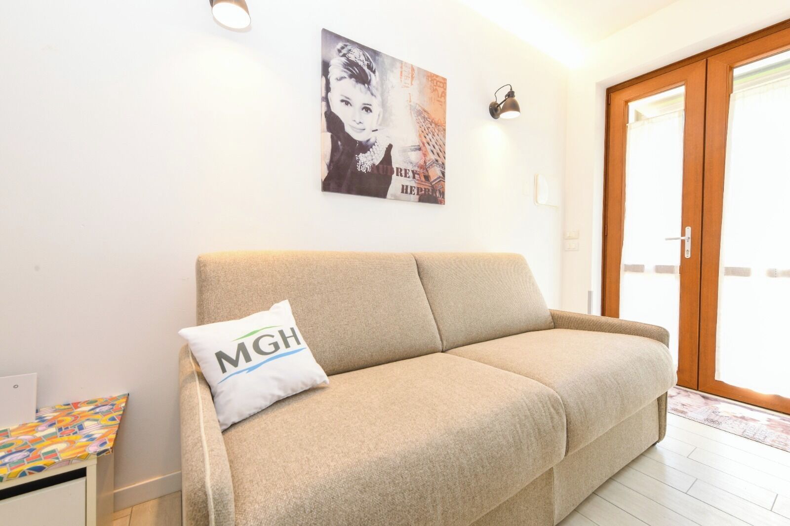 A beige sofa with a pillow labeled "MGH" in a room with a wooden door and a poster on the wall.