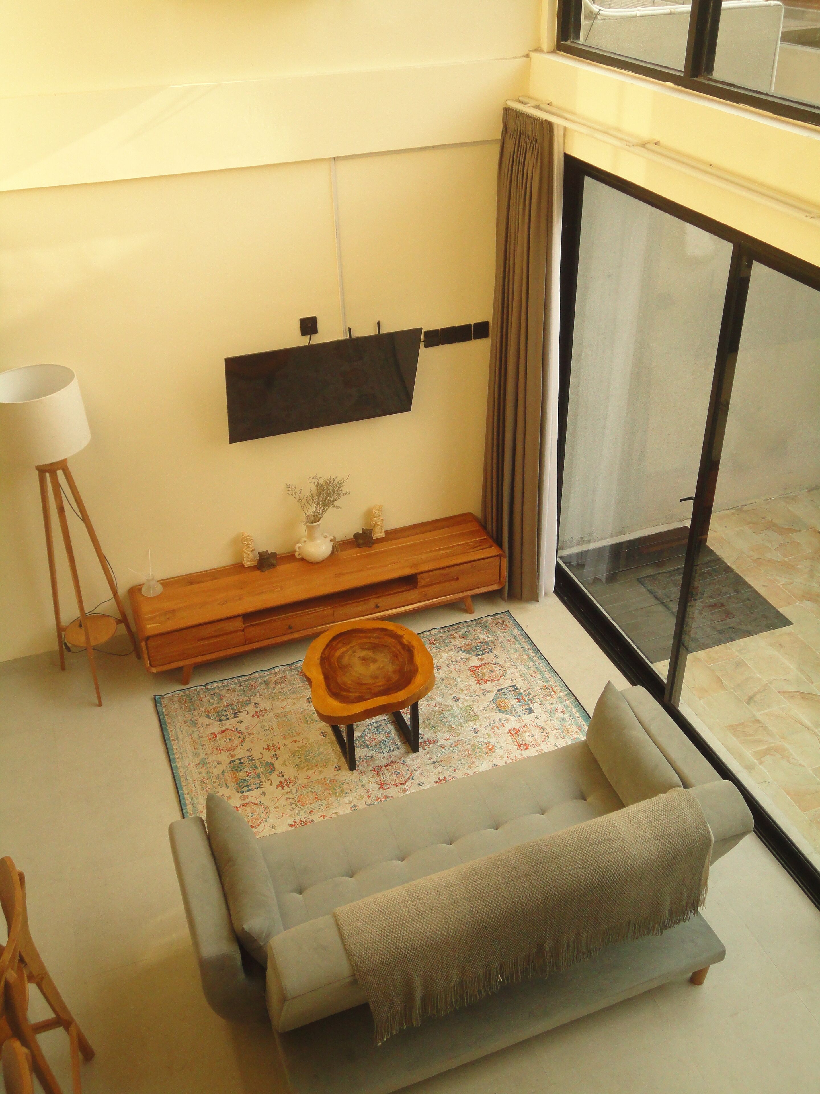 A living room with a sofa, a small wooden stool, a floor lamp, a flat-screen TV mounted on the wall, and a patterned rug.