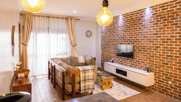 A cozy living room with a brick wall, a sofa, a coffee table, and a TV mounted on the wall. - Charming 2-bedroom Apartment in Bwebajja, Kampala (Entebbe)