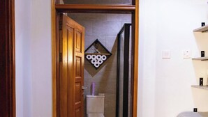 A narrow hallway with a wooden door leading to a bathroom. There is a wooden shoe rack, a pair of shoes on a mat, and a woven basket on the floor. - Charming 2-bedroom Apartment in Bwebajja, Kampala (Entebbe)
