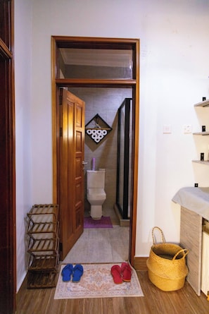 A narrow hallway with a wooden door leading to a bathroom. There is a wooden shoe rack, a pair of shoes on a mat, and a woven basket on the floor. - Charming 2-bedroom Apartment in Bwebajja, Kampala (Entebbe)
