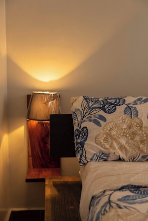 A neatly made bed with a blue and white floral bedspread, a wooden nightstand with a lamp, and a wall-mounted shelf. - Charming 2-bedroom Apartment in Bwebajja, Kampala (Entebbe)