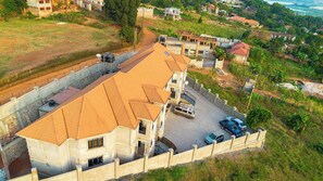 A multi-story building with a tiled roof, surrounded by a low wall and a parking area with several cars. - The Apartments Bwebajja (Entebbe)