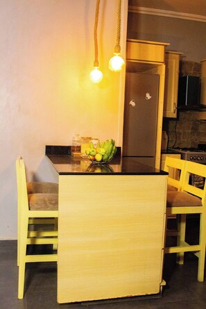 A compact kitchen with a wooden island, two chairs, a fruit bowl, and a hanging light fixture. - The Apartments Bwebajja (Entebbe)