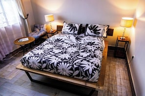 Ein Bett mit floralem Überwurf, zwei Nachttischlampen, ein kleiner runder Tisch und ein Sessel mit Kissen.