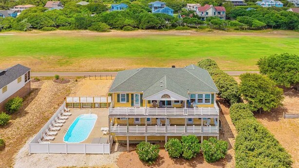 Una casa de dos pisos con piscina, rodeada de vegetación y otras casas al fondo.