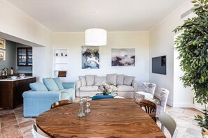 Ein modernes Wohnzimmer mit einem Holzespeisetisch, einem hellblauen Sofa, einem beigen Sofa und einer großen Grünpflanze.