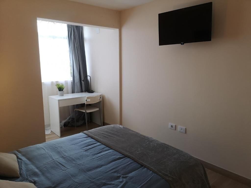 Een slaapkamer met een bed, een bureau, een stoel, een televisie en een raam met gordijnen.