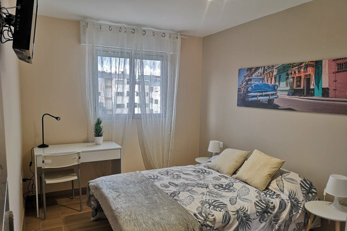 Een slaapkamer met een bed, een bureau, een stoel, een kamerplant en een schilderij aan de muur.
