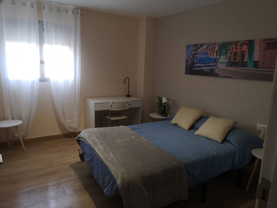 A bedroom with a bed, desk, chair, and a painting on the wall.