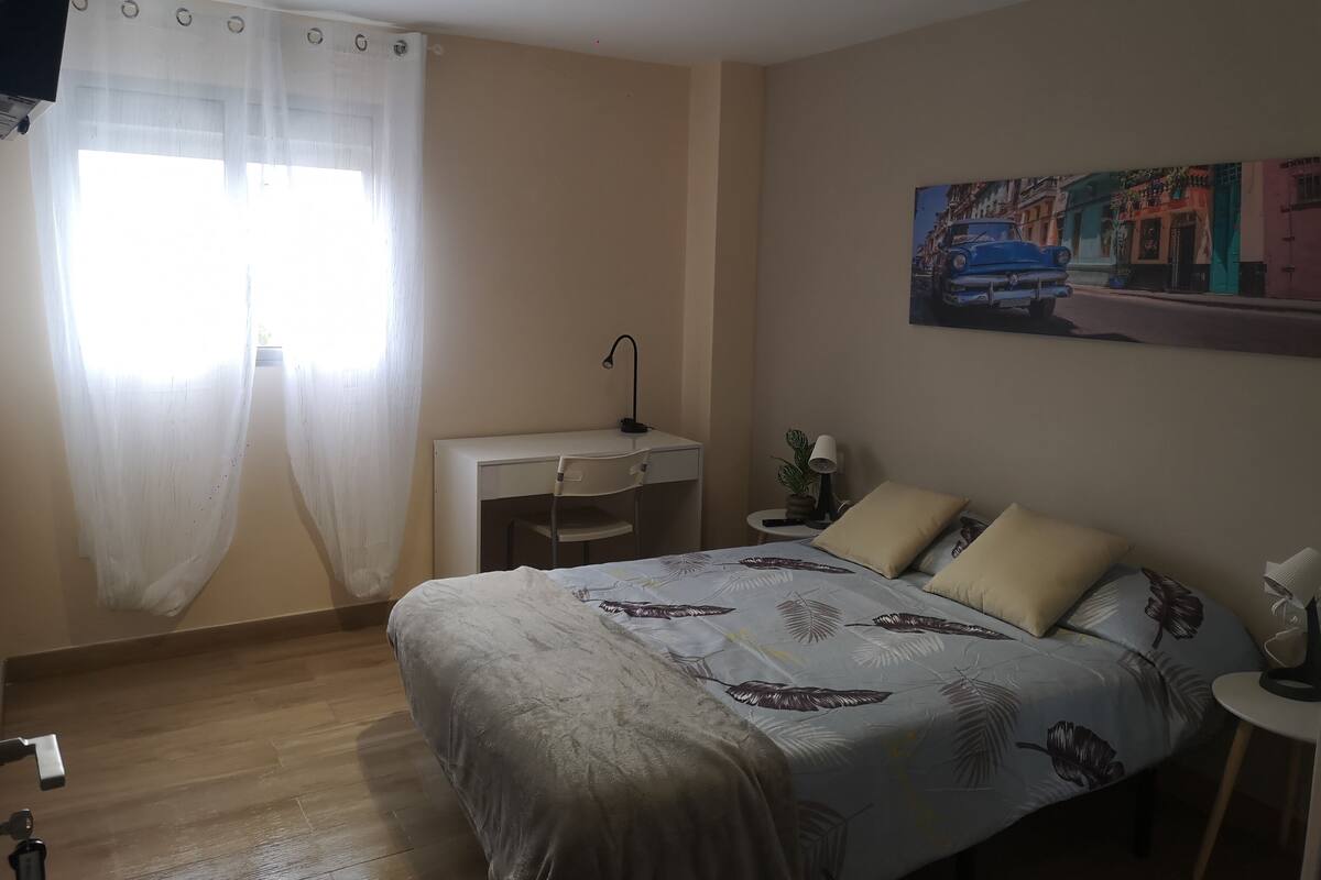 Een slaapkamer met een bed, een bureau en een stoel. Er is een raam met dunne gordijnen.