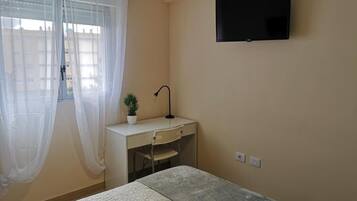A bedroom with a bed, a desk with a chair, a potted plant, a TV mounted on the wall, and a window with sheer curtains.