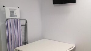 Quarto pequeno com uma mesa branca e quatro cadeiras. Há uma televisão na parede e uma cortina listrada.