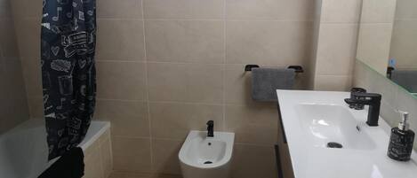 Un baño con bidé, un lavamanos blanco, un espejo y una ventana con cortina.