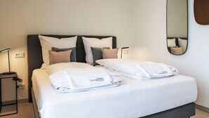 Ein ordentlich bezogenes Bett mit weißen Leinen und einem gefalteten Handtuch, eine Nachttischlampe und ein an der Wand befestigter Spiegel.