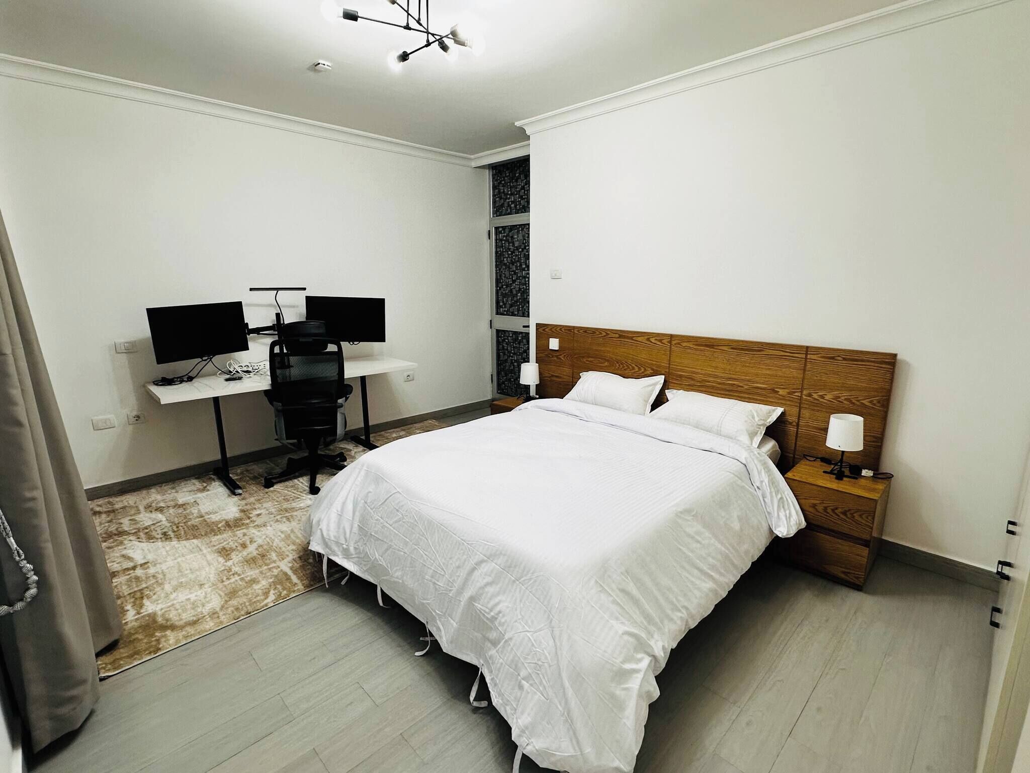 Een moderne slaapkamer met een groot bed, een bureau met twee monitoren en een nachtkastje met een lamp.