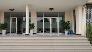 Entrada de un edificio con una escalera que conduce a una puerta de cristal, flanqueada por dos columnas y plantas en macetas.