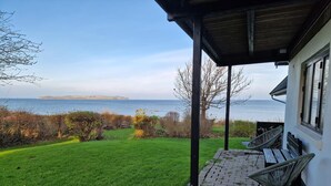 A covered patio with a view of the sea, a grassy lawn, and a white building. - Waterfront holiday for multiple generations (Føllenslev)
