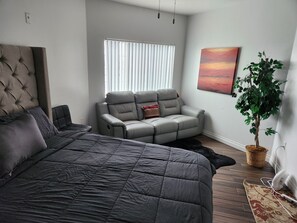 Een moderne slaapkamer met een groot bed, een grijze relaxfauteuil, een potplant en een schilderij aan de muur.