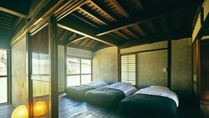 Una habitación tradicional japonesa con suelo de tatami, techo bajo con vigas de madera y una puerta corrediza estilo shoji. Se observan tres paneles de tatami colocados en fila, con almohadones y un bajo catamarán de madera.