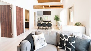 A modern living room with a grey sofa, wooden ceiling beams, and a dining area in the background.