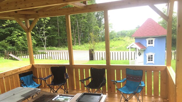 A covered outdoor area with a brick fire pit table, several folding chairs, and a view of a blue house with a red roof in the background.