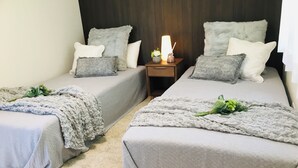 A hotel room with two single beds, each with a grey bedspread and pillows, a small bedside table with a lamp, and a decorative plant on each bed.