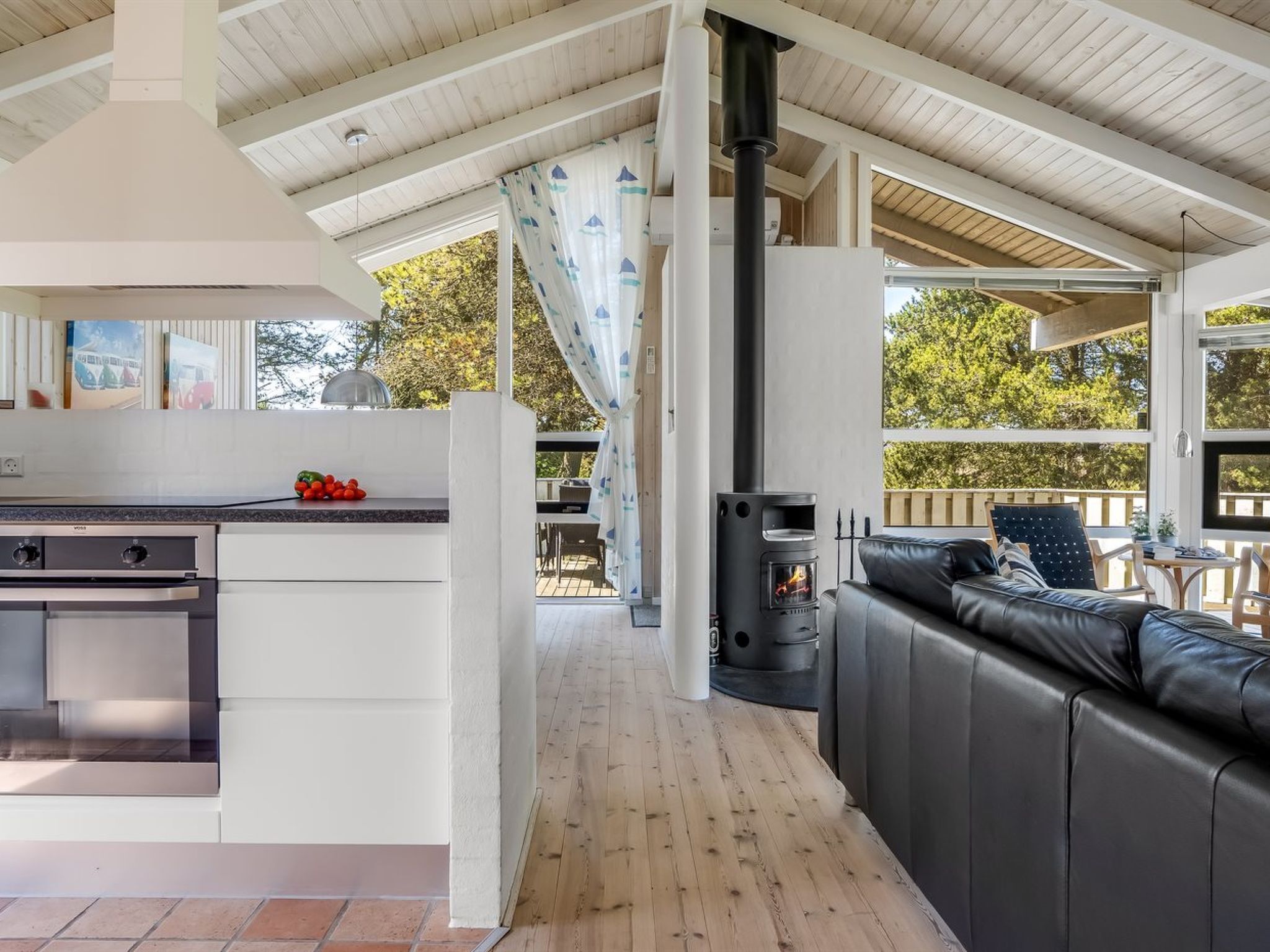 A modern kitchen with a built-in stove, a dining area with a black leather couch, and a large window overlooking a deck.