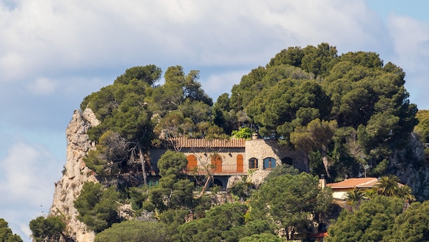 Un bâtiment en pierre, doté de fenêtres en arc de cercle et d’un toit recouvert de tuiles rouges, entouré d’une forêt dense et d’une falaise rocheuse.