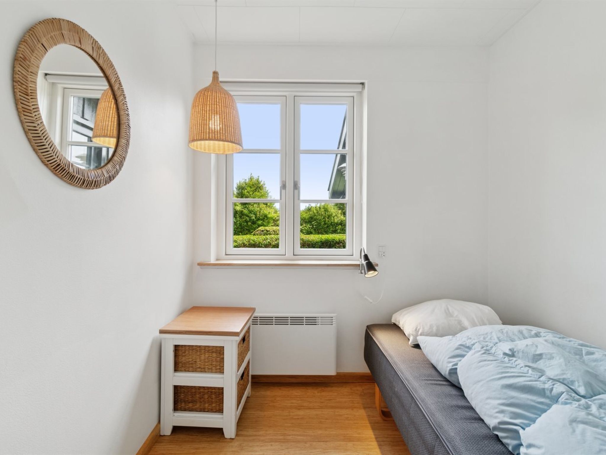 A bedroom with a bed, a bedside table, a mirror, a window with a view of greenery, and a hanging light fixture.