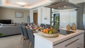 A modern kitchen with a central island, dining area with chairs and a table set for a meal, and a living area with a sofa and TV. - IMPERIUM LUXURY VILLAS (Chania)