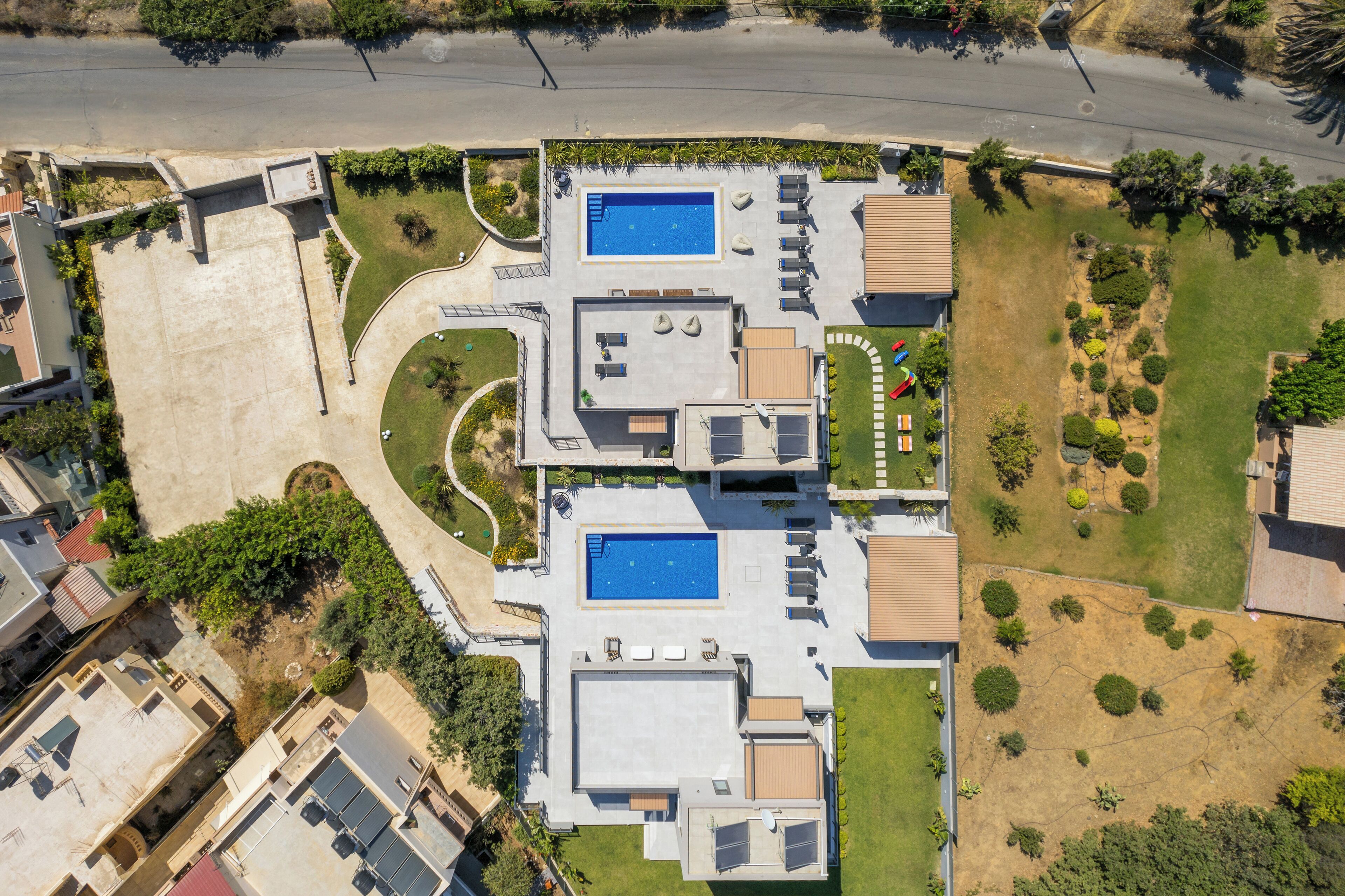 Vista aérea de una propiedad residencial con dos piscinas, un jardín ajardinado y una carretera al fondo.