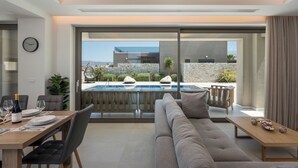 A modern living room with a sofa, dining table, and chairs, overlooking a pool and outdoor area with plants and a stone wall. - IMPERIUM LUXURY VILLAS (Chania)