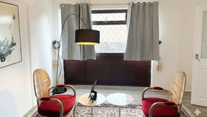 Een kamer met twee rieten stoelen met rode kussens, een kleine ronde tafel, een vloerlamp en een raam met gordijnen.
