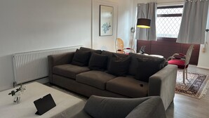 Een moderne woonkamer met een hoekbank, een klein tafeltje met een tablet, een radiator, een raam met gordijnen en een vloerlamp.