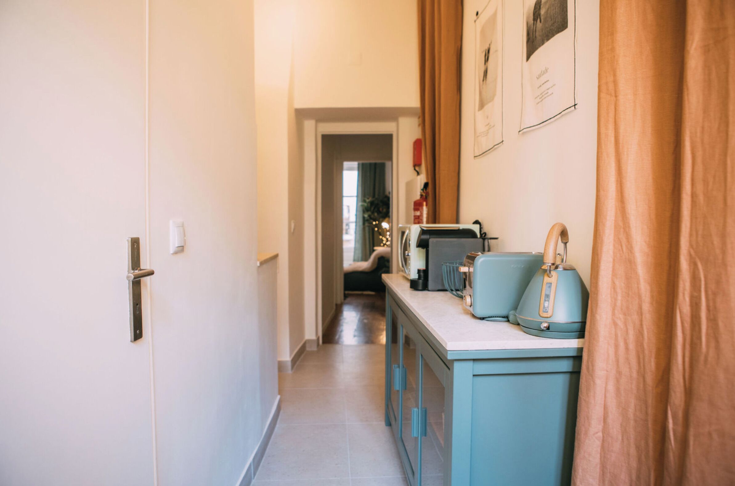 A narrow hallway with a door on the left, a kitchen area with a blue cabinet and a kettle on the right, and a curtain partially covering the right side.
