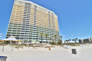 A high-rise hotel with multiple balconies, a beach area with palm trees, and a clear blue sky. - Boardwalk Beach Resort Condo | 2BR Sleeps 6 | Direct Beach Access (Panama City Beach)