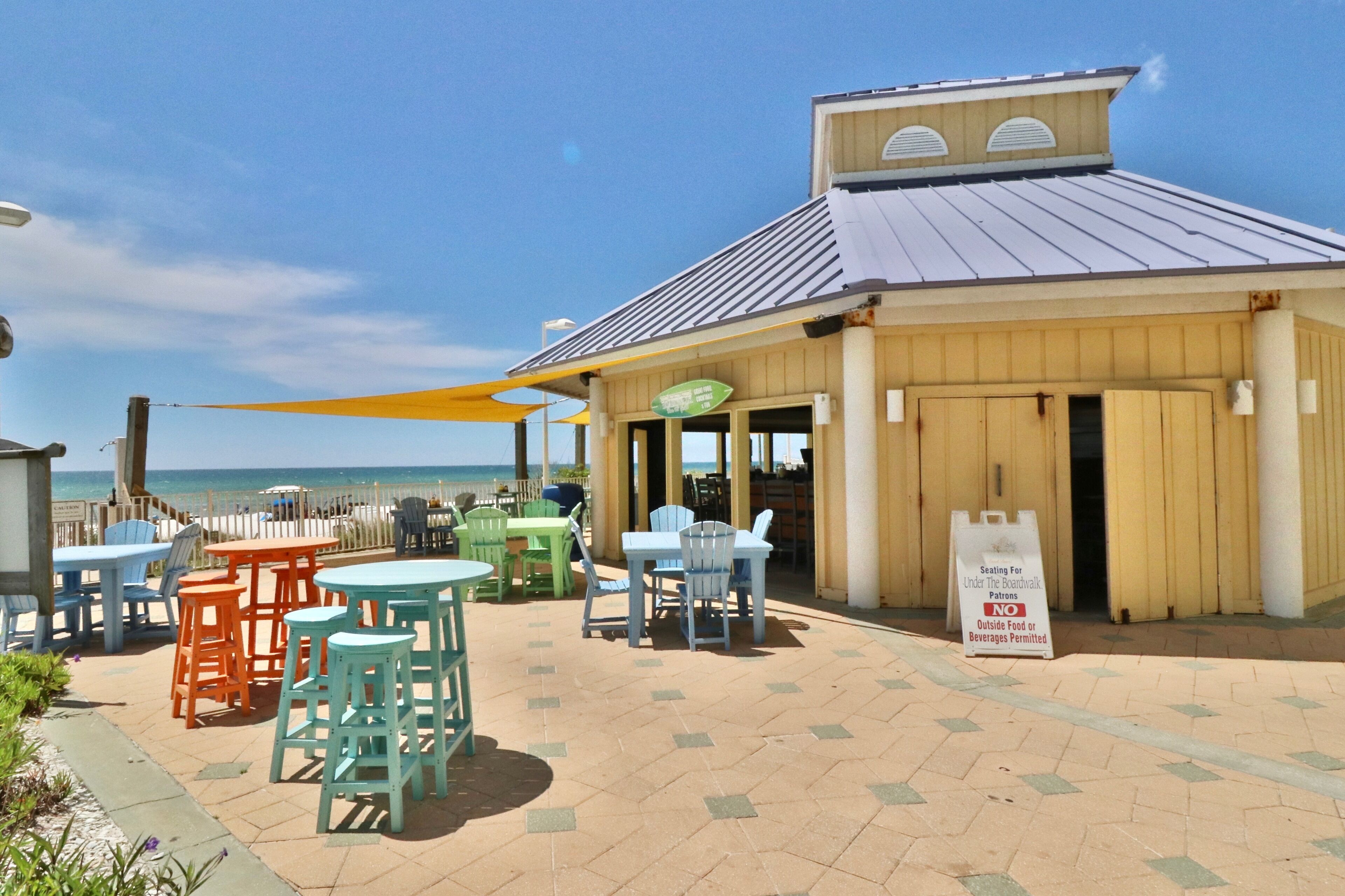 Un espace extérieur aménagé avec des tables et des chaises, un panneau indiquant qu’il est interdit d’apporter de la nourriture ou des boissons de l’extérieur, et une vue sur la plage.