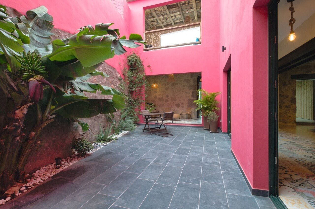 A courtyard with a pink wall, a stone wall, and a tiled floor. There is a table and chairs in the center, surrounded by plants and a potted plant.