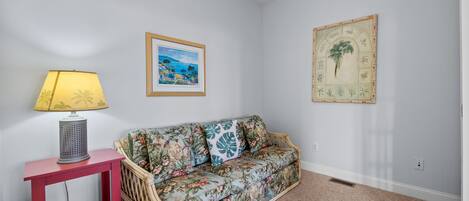 Ein Zimmer mit einer sofaartigen Sitzgelegenheit mit Blumenmuster, einem kleinen roten Beistelltischchen mit Lampe und zwei gerahmten Kunstwerken an den Wänden.