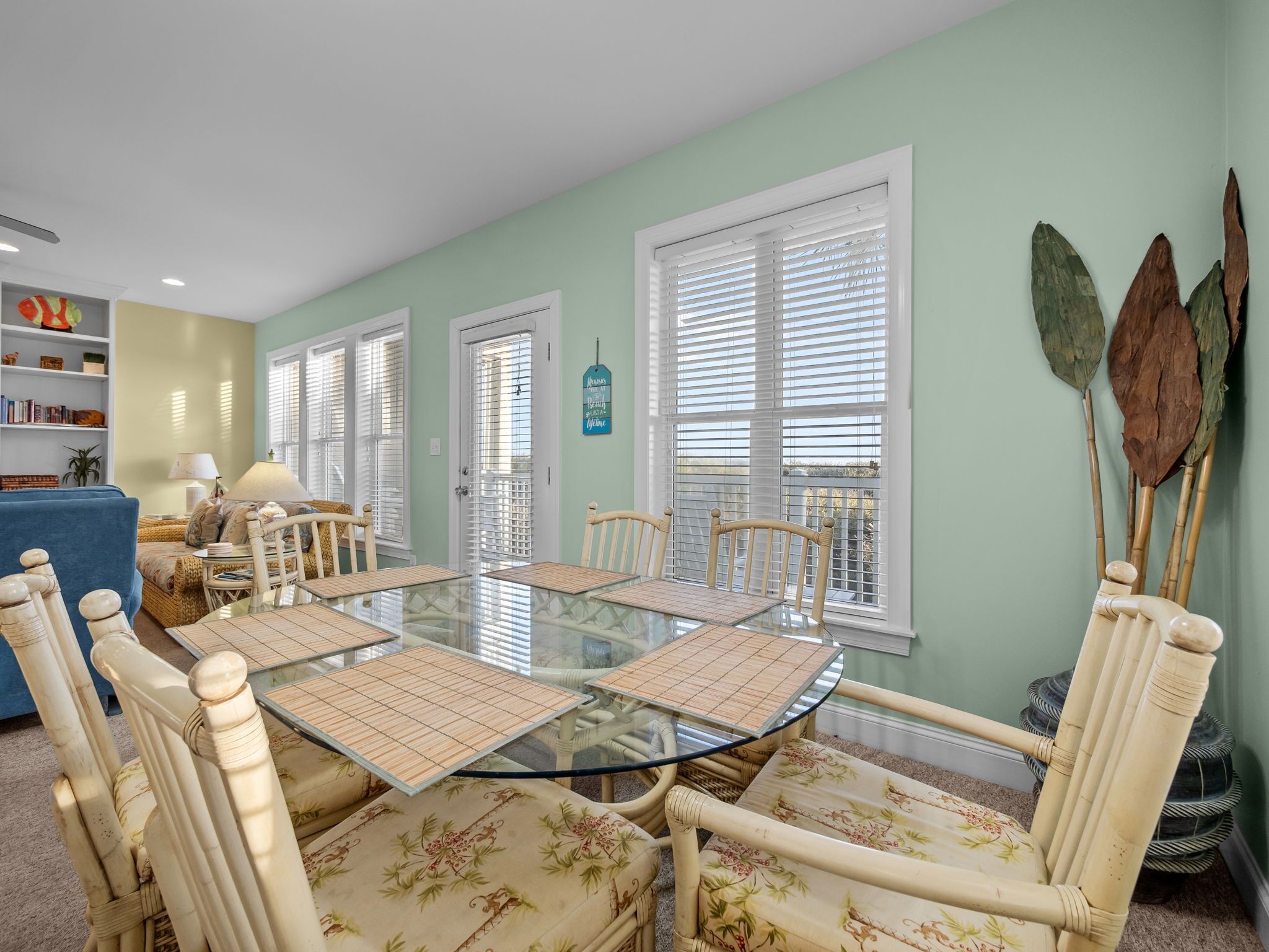 Un coin repas avec une table à plateau de verre entourée de chaises à motifs floraux, de grandes fenêtres équipées de stores et une bibliothèque contenant divers objets.