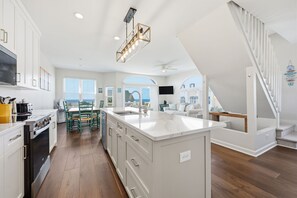 A modern kitchen with white cabinetry, a central island, and a dining area with green chairs. - Beach Allure West | Pet-Friendly Oceanfront Duplex (Emerald Isle)