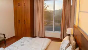 A bedroom with a bed, a window with curtains, a wooden wardrobe, and a nightstand with a lamp.