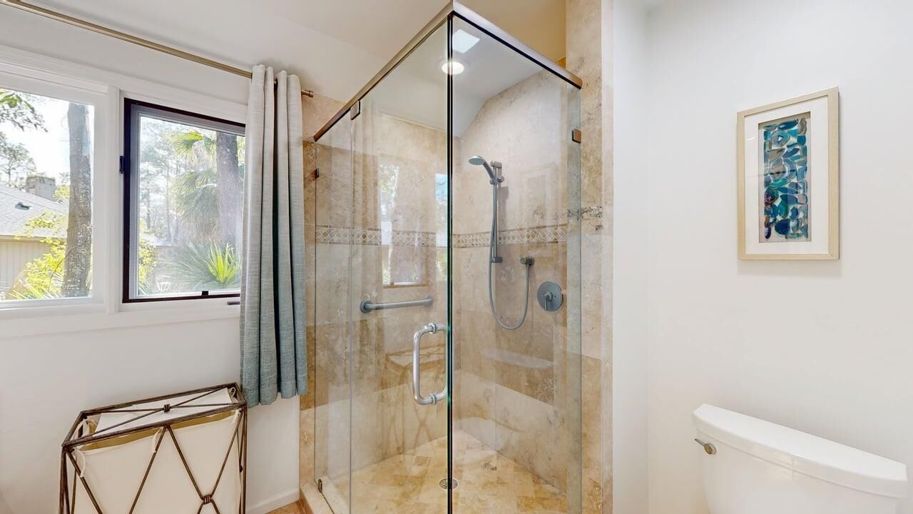 Un baño con ducha envuelta en vidrio, un inodoro, una ventana con cortinas y una obra de arte enmarcada en la pared.