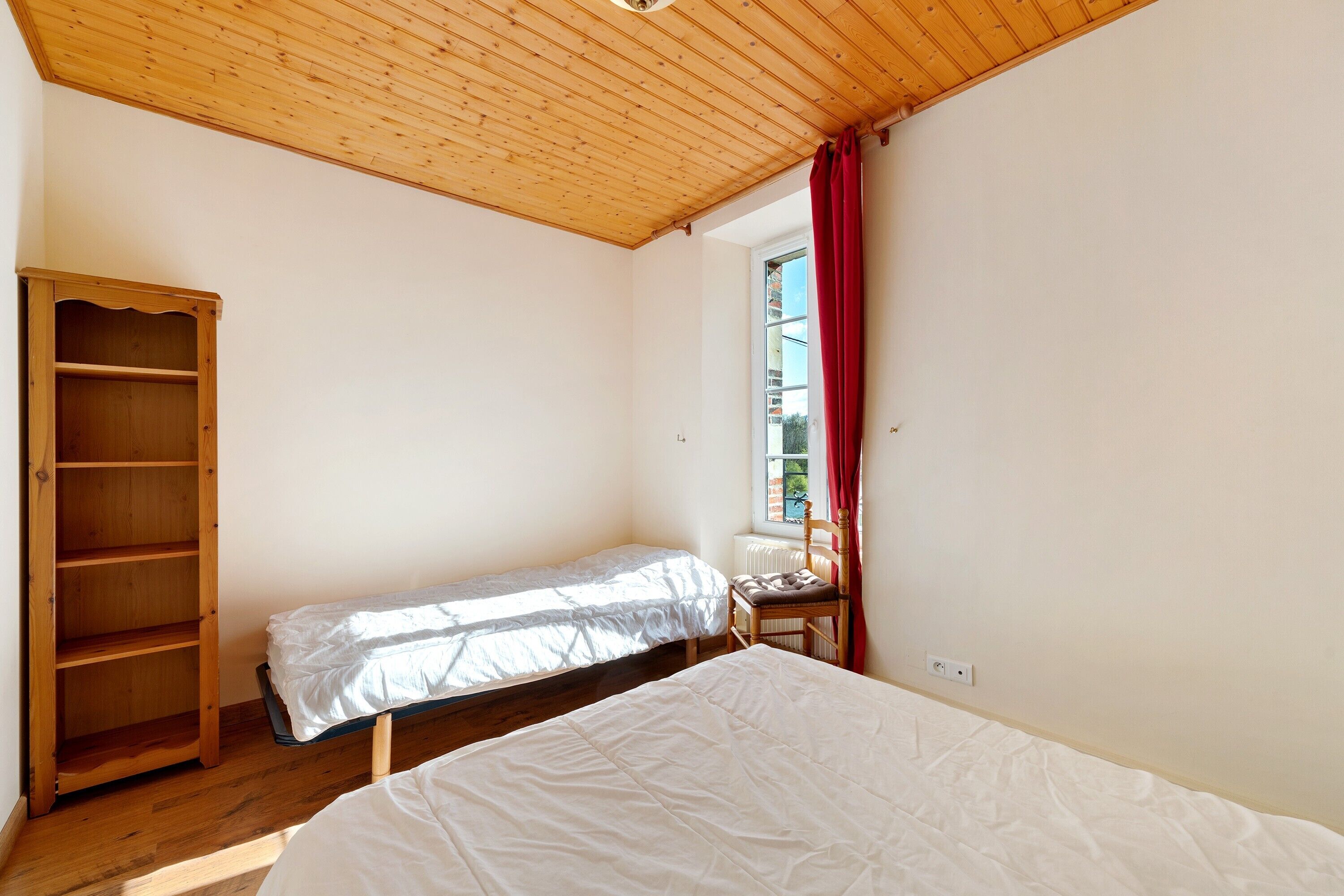 A room with a wooden ceiling, a bed, a bench, and a window with red curtains.