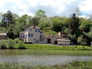 A two-story house with a balcony, a small attached structure, and a pond in the foreground.