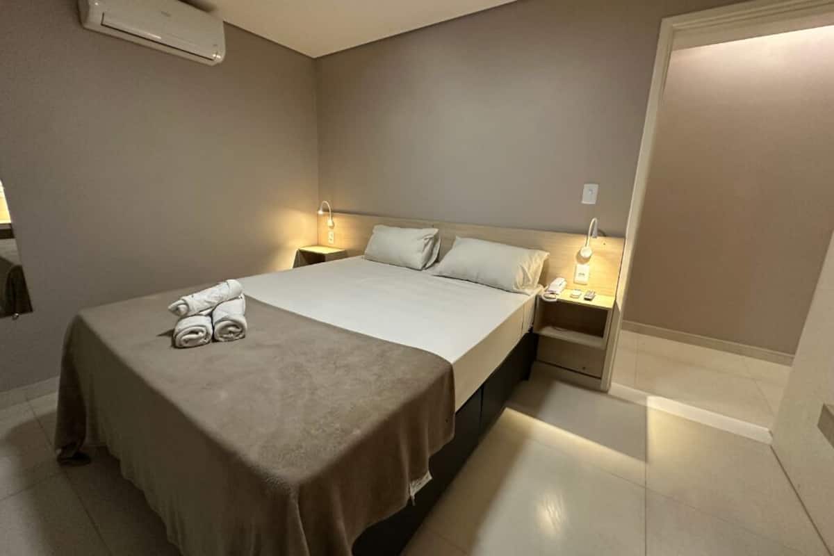 A hotel room with a bed, two bedside tables, a nightstand, and a wall-mounted air conditioner.